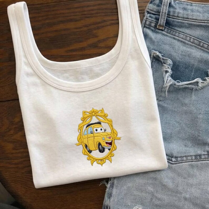 Op de afbeelding: Witte tanktop met een gele cartoon auto geborduurd op de voorkant, omlijst door een goudkleurige decoratieve rand. De tanktop wordt gecombineerd met een spijkerbroek short. De algehele uitstraling is casual en speels.