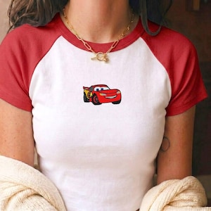 May include: A white raglan t-shirt with short red sleeves and a red crew neckline. The centre of the chest features a small, embroidered graphic of a red race car with blue eyes, yellow lightning bolt accents, and a smiling mouth on the front bumper.