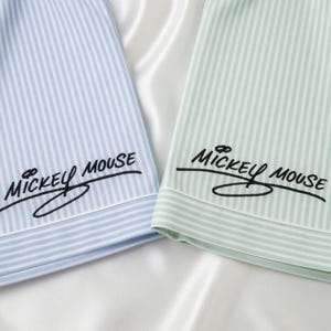 May include: Close-up of two pajama tops. One is light blue with white vertical stripes, the other is light green with white stripes. Both have the embroidered text "Mickey Mouse" in black script.