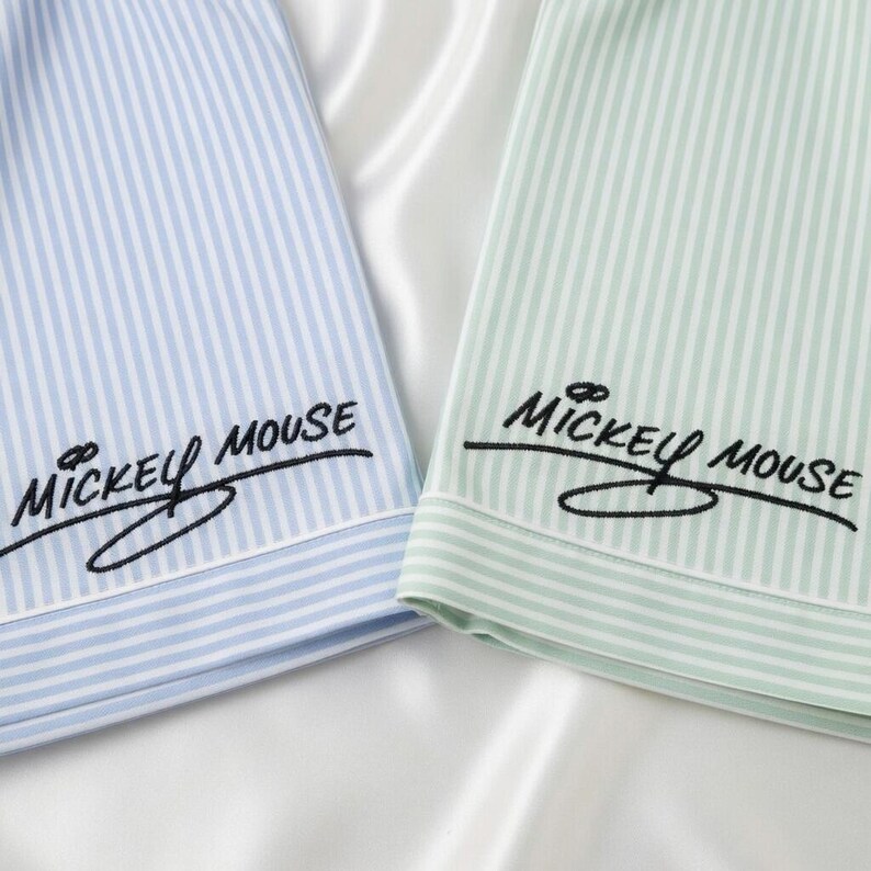 May include: Close-up of two pajama tops. One is light blue with white vertical stripes, the other is light green with white stripes. Both have the embroidered text "Mickey Mouse" in black script.