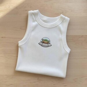 K&ouml;nnte beinhalten: Wei&szlig;er &auml;rmelloser Baby-Body mit rundem Halsausschnitt. Der Body hat ein gesticktes Bild eines gr&uuml;nen Alien-Charakters in einem Raumschiff. Der Au&szlig;erirdische befindet sich in einer durchsichtigen Kuppel. Das Raumschiff ist grau und braun.