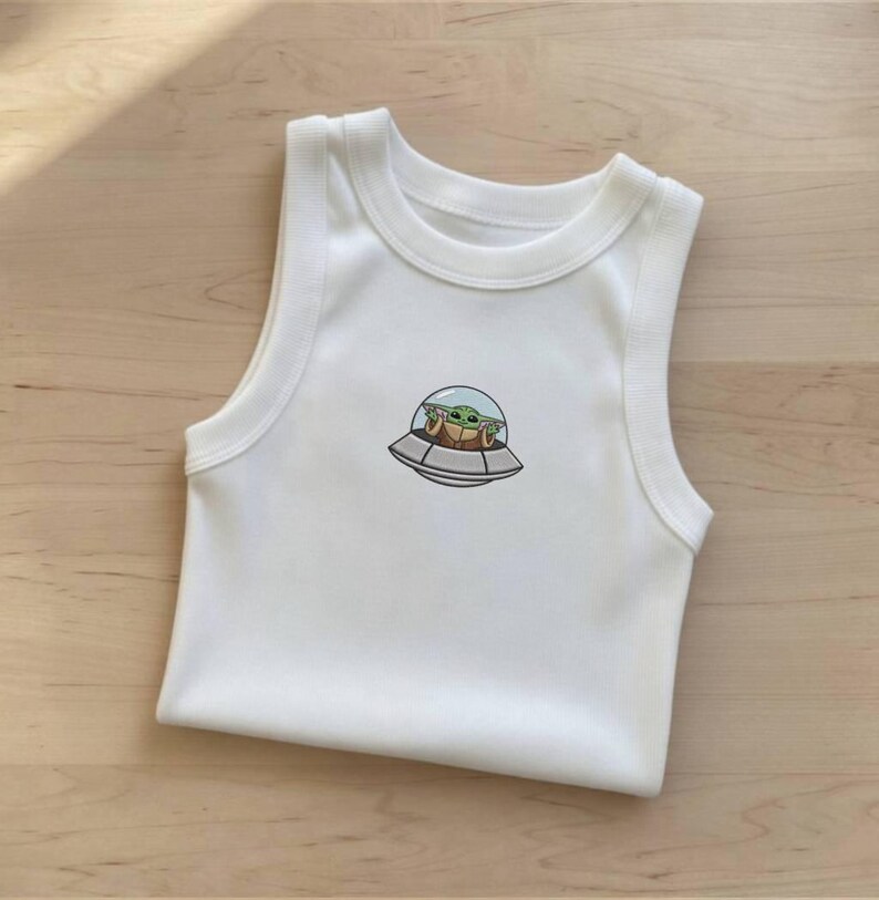 K&ouml;nnte beinhalten: Wei&szlig;er &auml;rmelloser Baby-Body mit rundem Halsausschnitt. Der Body hat ein gesticktes Bild eines gr&uuml;nen Alien-Charakters in einem Raumschiff. Der Au&szlig;erirdische befindet sich in einer durchsichtigen Kuppel. Das Raumschiff ist grau und braun.