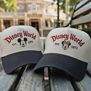 Op de afbeelding: Twee baseballcaps met de woorden "Disney World" in rode letters. Eén cap heeft Mickey Mouse, de andere Minnie Mouse, beide met "EST. 1971". De caps hebben een crèmekleurige kroon en een donkerblauwe klep.