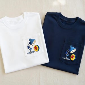 Op de afbeelding: Twee T-shirts, &eacute;&eacute;n wit en &eacute;&eacute;n marineblauw, elk met een zakje met een geborduurd ontwerp. Het ontwerp toont een blauwe bureaulamp en een gele cirkel met een rode ster. De shirts zijn gemaakt van zacht materiaal, waarschijnlijk katoen.