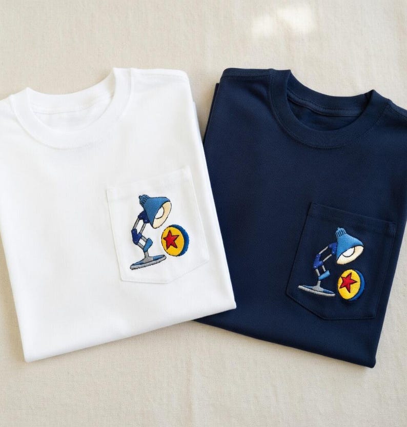 Op de afbeelding: Twee T-shirts, &eacute;&eacute;n wit en &eacute;&eacute;n marineblauw, elk met een zakje met een geborduurd ontwerp. Het ontwerp toont een blauwe bureaulamp en een gele cirkel met een rode ster. De shirts zijn gemaakt van zacht materiaal, waarschijnlijk katoen.
