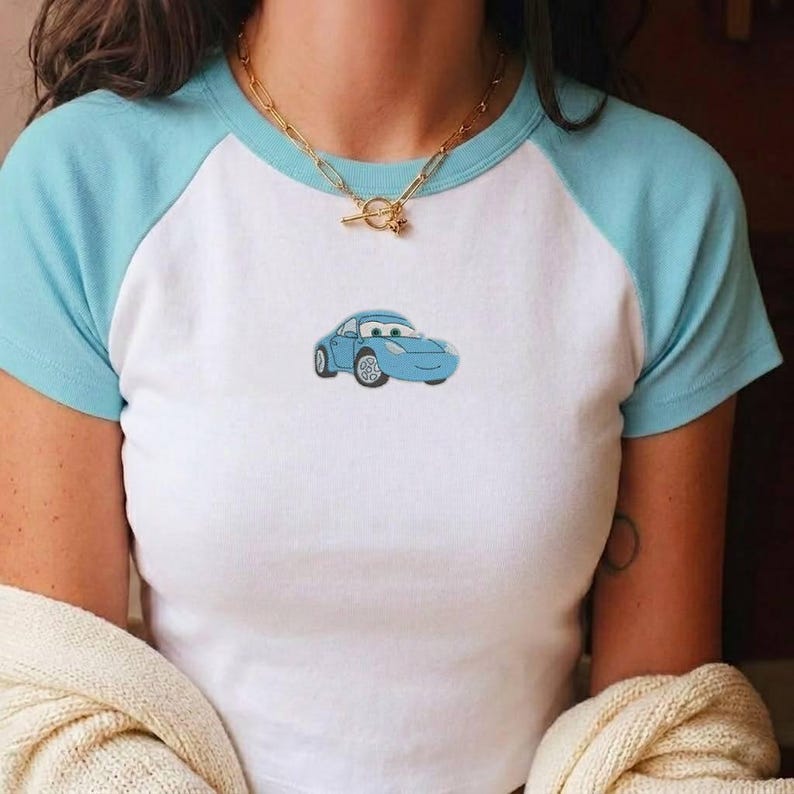May include: A white and light blue raglan t-shirt featuring a small embroidered blue car graphic on the centre chest. The shirt has a round neckline and is paired with a gold-toned chain necklace with a toggle clasp closure at the front.