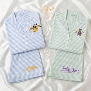 Op de afbeelding: Twee sets gestreepte pyjama's. De ene set is lichtblauw met Pluto geborduurd op de zak en de shorts. De andere set is lichtgroen met Daisy Duck geborduurd op de zak en de shorts. Beide sets hebben witte biezen.