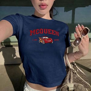 May include: Navy blue crop top with the word "MCQUEEN" in red above a cartoon image of a red race car. The text "I am speed" is printed below the car. The shirt is a casual style.