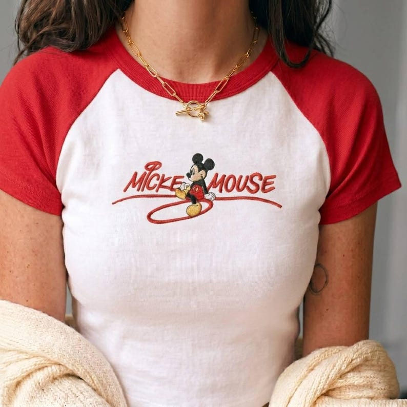 Op de afbeelding: Een wit raglan t-shirt met rode mouwen en een rode kraag. Het midden van het shirt is voorzien van een geborduurde afbeelding van Mickey Mouse in zijn klassieke rode broek en gele schoenen, met de tekst Mickey Mouse in rood cursief schrift.