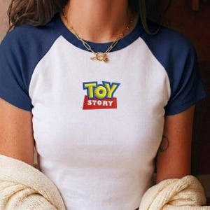 May include: A white short-sleeved t-shirt with dark blue raglan sleeves and a round neckline. The center of the chest features an embroidered Toy Story logo with yellow letters on a blue background and white letters on a red rectangular base.
