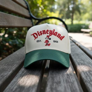 May include: A white and green baseball cap with the word "Disneyland" embroidered in red script. The cap features a Minnie Mouse graphic and the text "EST. 1955". The cap is sitting on a wooden bench.