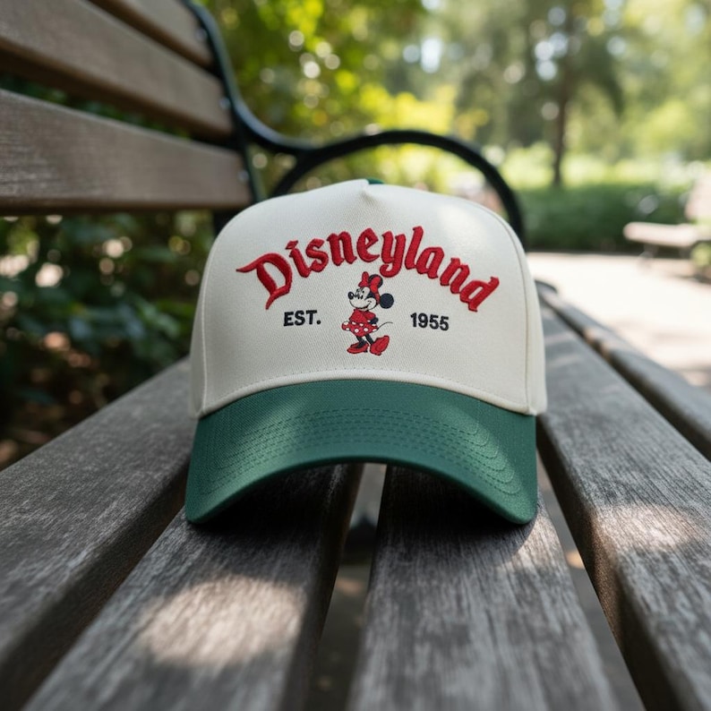 May include: A white and green baseball cap with the word "Disneyland" embroidered in red script. The cap features a Minnie Mouse graphic and the text "EST. 1955". The cap is sitting on a wooden bench.