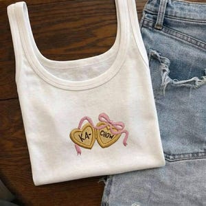 K&ouml;nnte beinhalten: Wei&szlig;es Tanktop mit Rundhalsausschnitt und gestickten herzf&ouml;rmigen Keksen. Die Kekse sind goldfarben mit rosa Akzenten, die Texte "KA" und "CHOW" sind auf die Kekse gestickt. Das Tanktop wird mit Jeansshorts kombiniert.
