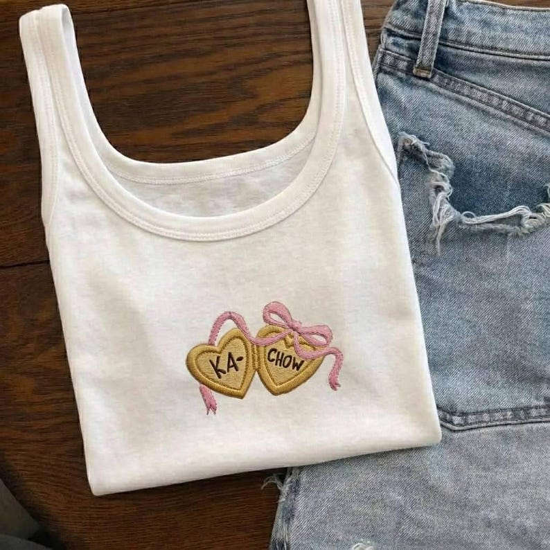 K&ouml;nnte beinhalten: Wei&szlig;es Tanktop mit Rundhalsausschnitt und gestickten herzf&ouml;rmigen Keksen. Die Kekse sind goldfarben mit rosa Akzenten, die Texte "KA" und "CHOW" sind auf die Kekse gestickt. Das Tanktop wird mit Jeansshorts kombiniert.