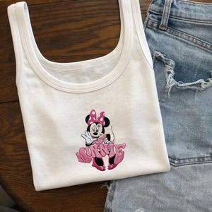 Może przedstawiać: Biały top na ramiączkach z r&oacute;żowo-czarnym motywem Myszki Minnie. Postać z kresk&oacute;wki ma na sobie r&oacute;żową sukienkę i kokardę. Top na ramiączkach jest połączony z dżinsowymi szortami.
