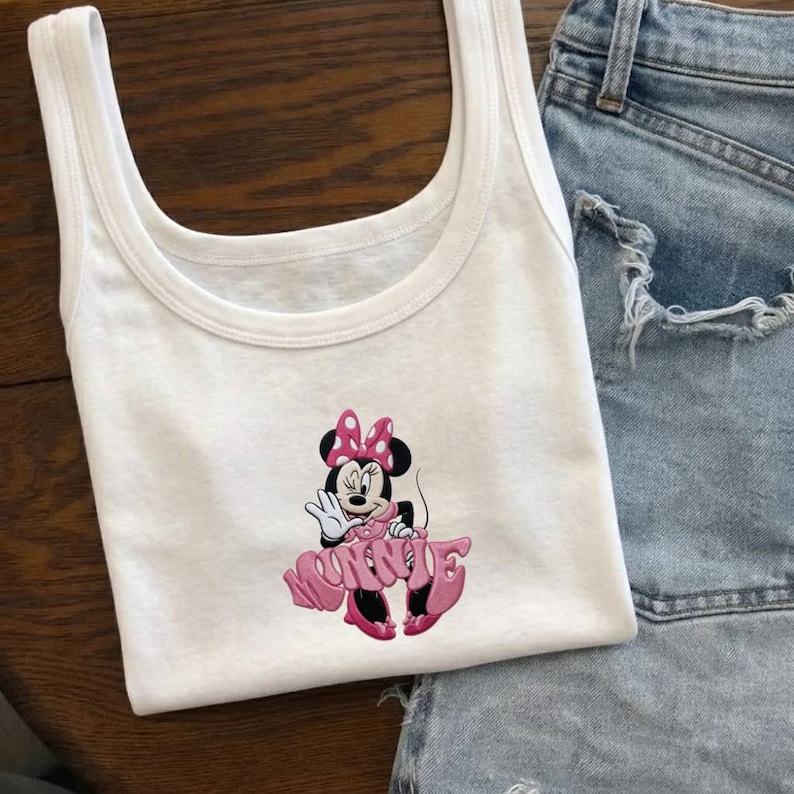 Może przedstawiać: Biały top na ramiączkach z r&oacute;żowo-czarnym motywem Myszki Minnie. Postać z kresk&oacute;wki ma na sobie r&oacute;żową sukienkę i kokardę. Top na ramiączkach jest połączony z dżinsowymi szortami.