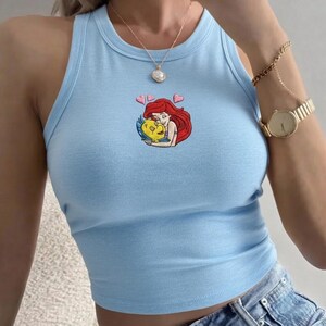 May include: Pale blue vest top with a round neck. The top has an embroidered design of a red-haired woman hugging a yellow fish, with two pink hearts above. The top is made of a soft material.