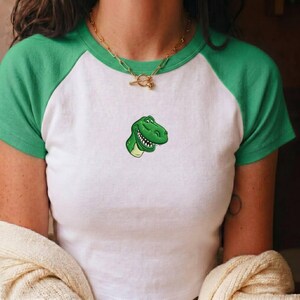 May include: A white and green raglan t-shirt featuring a small, centered graphic of a smiling green dinosaur head. The shirt has a round neckline and short sleeves. A gold-toned chain necklace with a toggle clasp is worn around the neck.