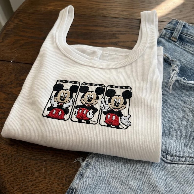 Puede incluir: Camiseta sin mangas blanca acanalada con un dise&ntilde;o bordado de Mickey Mouse en tres poses diferentes. El personaje de dibujos animados lleva pantalones cortos rojos y guantes blancos. La camiseta sin mangas se combina con pantalones cortos vaqueros azul claro.