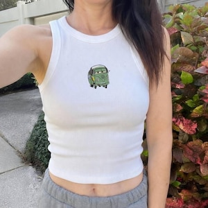 May include: White ribbed tank top with a small embroidered cartoon car design in green, yellow, and black. The sleeveless top has a round neckline.