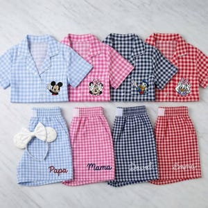 May include: Matching pajama sets in blue, pink, navy, and red gingham patterns. Each set includes a short-sleeved button-down shirt with a cartoon character and matching shorts with embroidered names. A white bow headband is included.