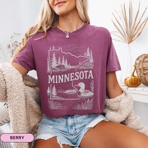 Op de afbeelding: Een paars, gecropped T-shirt met een witte omtrek van de staat Minnesota, een zeemeeuw en bomen. De tekst "MINNESOTA" is op het shirt gedrukt.