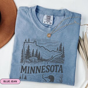Op de afbeelding: Een jeansblauw T-shirt met een zwart-witte afbeelding van de staat Minnesota met een duiker en de tekst "Minnesota".