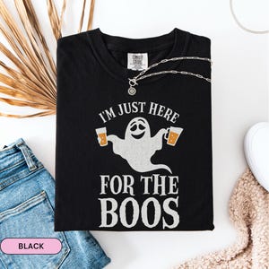 Op de afbeelding: Zwart T-shirt met de tekst "I'M JUST HERE FOR THE BOOS" en een cartoon spook met twee bierpullen. Een zilveren ketting is over het shirt gedrapeerd. Het shirt wordt gepresenteerd op een witte ondergrond met een spijkerbroek en een beige trui.