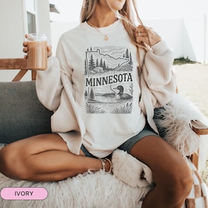 Op de afbeelding: Een wit sweatshirt met een zwart-witte afbeelding van de staat Minnesota met een zeemeeuw op een meer. De tekst "MINNESOTA" staat op het sweatshirt gedrukt. Het sweatshirt wordt gedragen door iemand die op een bank zit.