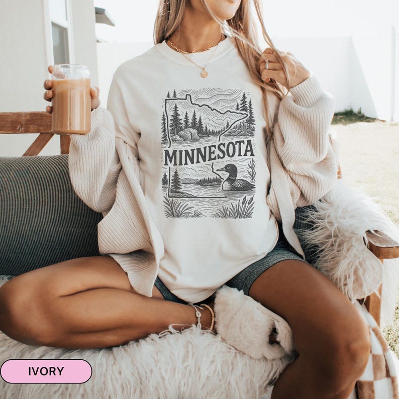 Op de afbeelding: Een wit sweatshirt met een zwart-witte afbeelding van de staat Minnesota met een zeemeeuw op een meer. De tekst "MINNESOTA" staat op het sweatshirt gedrukt. Het sweatshirt wordt gedragen door iemand die op een bank zit.