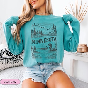 Op de afbeelding: Zeegroen sweatshirt met lange mouwen met een zwarte afbeelding van de staat Minnesota, bomen, een meer en een ijsvogel. Het woord "MINNESOTA" is vetgedrukt onder de afbeelding gedrukt. De sweatshirt is gemaakt van een zacht materiaal.