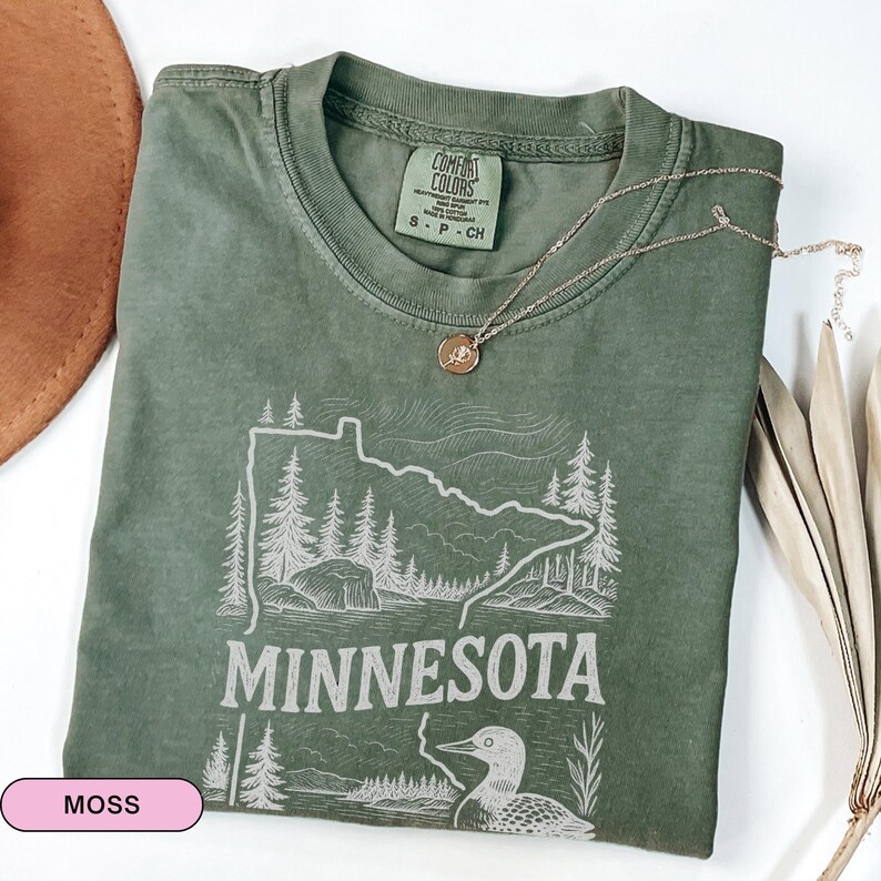 Op de afbeelding: Een mosgroen T-shirt met een witte omtrek van de staat Minnesota, een zeemeeuw en het woord "Minnesota". Het shirt heeft een label met de tekst "Comfort Colors" en "S - P - CH".