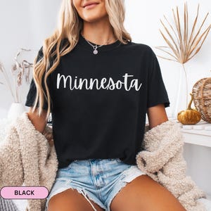 Op de afbeelding: Zwart T-shirt met het woord "Minnesota" in witte letters. Het model draagt een crèmekleurig vest en een spijkerbroek. Een kleine pompoen en een decoratieve droge plant staan op de achtergrond.