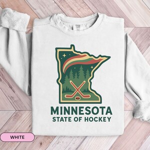Op de afbeelding: Wit sweatshirt met een groene omtrek van de staat Minnesota, een bos en hockey sticks grafisch. De tekst "MINNESOTA STATE OF HOCKEY" is eronder gedrukt. Het sweatshirt is gemaakt van zacht materiaal.