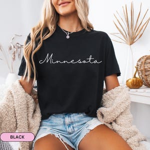 Op de afbeelding: Zwart T-shirt met het woord "Minnesota" in witte script. Het shirt heeft een ronde hals. De afbeelding toont een persoon die het shirt draagt met een lichtgekleurde jas en een blauwe spijkerbroek.
