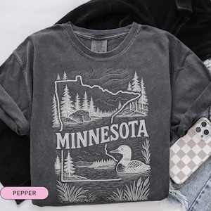 Op de afbeelding: Een donkergrijs T-shirt met een witte omtrek van de staat Minnesota en een zeemeeuw. De tekst "Minnesota" is in wit onder de omtrek gedrukt.