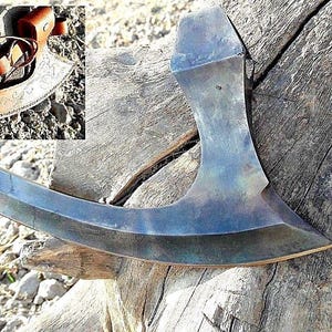 Könnte beinhalten: Eine gebogene, handgeschmiedete Axt mit einer dunkelblauen und grauen Patina. Die Axt liegt auf einem Stück verwittertem Holz.