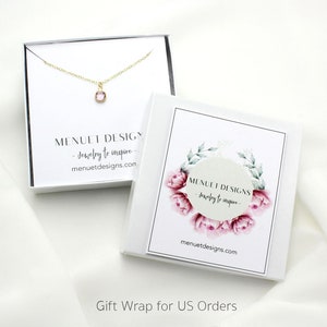 May include: A white gift box with a pink floral design and the text "MENUET DESIGNS - Jewelry to inspire - menuetdesigns.com". The box is open and a gold necklace with a small pink gemstone pendant is visible inside.
