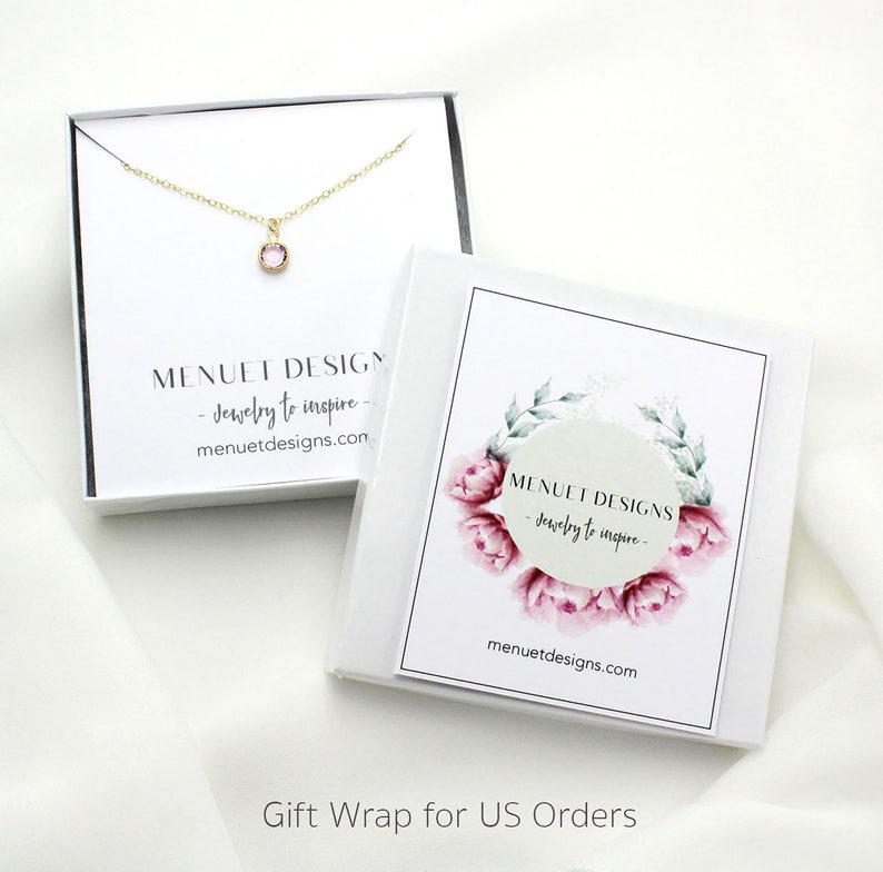 May include: A white gift box with a pink floral design and the text "MENUET DESIGNS - Jewelry to inspire - menuetdesigns.com". The box is open and a gold necklace with a small pink gemstone pendant is visible inside.