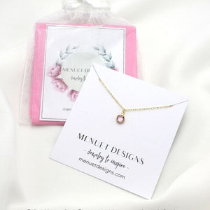 May include: A delicate gold necklace with a small pink gemstone pendant. The necklace is packaged in a white gift box with a pink tissue paper lining. The box is labeled with the text "MENUET DESIGNS - Jewelry to inspire - menuetdesigns.com".