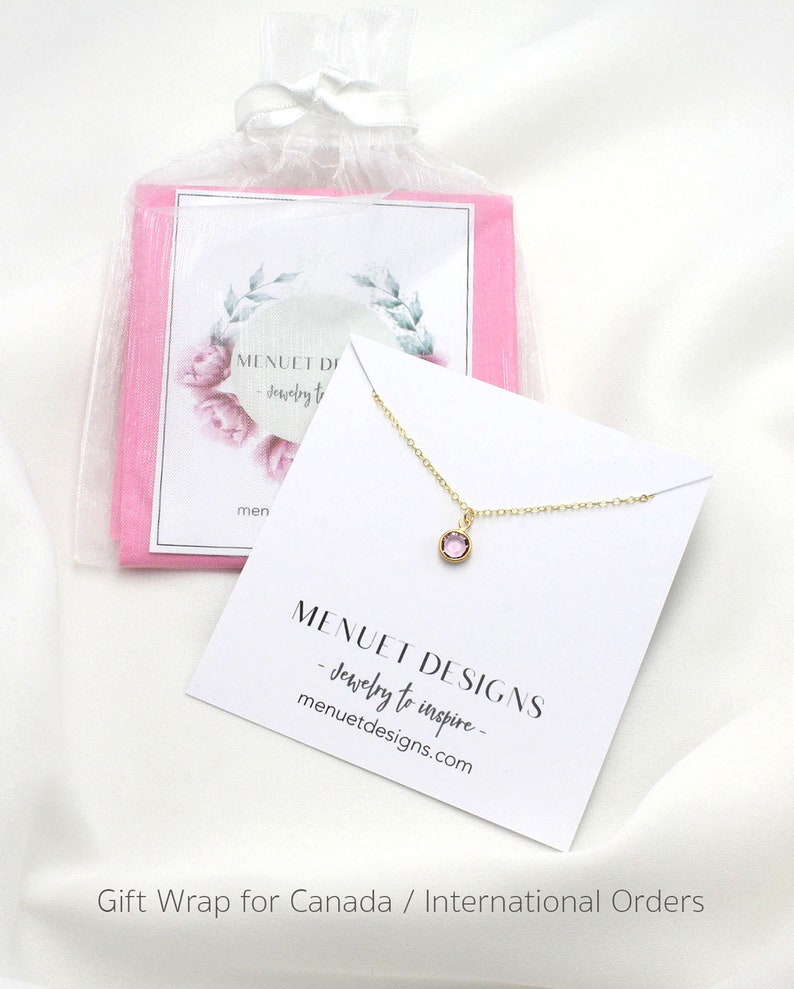 May include: A delicate gold necklace with a small pink gemstone pendant. The necklace is packaged in a white gift box with a pink tissue paper lining. The box is labeled with the text "MENUET DESIGNS - Jewelry to inspire - menuetdesigns.com".