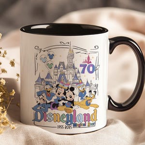 May include: White ceramic mug with a black interior and handle. The mug features a colorful illustration of Disneyland characters and the castle, with the text "Disneyland" and "70". The mug also has the text "1955-2025 Magical Years".