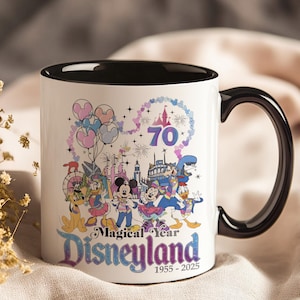 May include: White ceramic mug with a black interior and handle. The mug features a colorful graphic of Disney characters, fireworks, and the text "Magical Year Disneyland 1955-2025" celebrating its 70th anniversary.