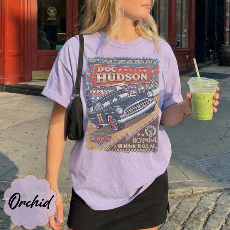 May include: A light purple short-sleeve t-shirt featuring a graphic print of the character Doc Hudson from the movie Cars. The design includes the text Doc Hudson, Radiator Springs Saturday Night Special Race, and Winner Takes All.