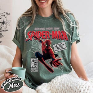 Op de afbeelding: Een mosgroen t-shirt met een Spider-Man stripboekontwerp. De afbeelding bevat de woorden "BRAND NEW DAY" en "SPIDER-MAN" in het rood, samen met een illustratie van Spider-Man en stripboektekst. Het shirt heeft ook de woorden "COMFORT COLORS" en "Moss".