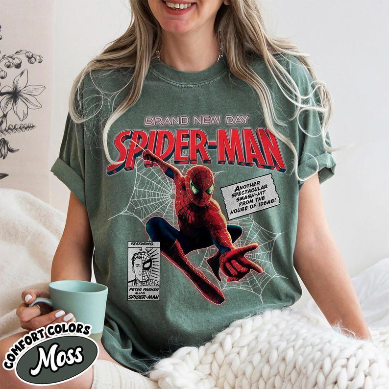 Op de afbeelding: Een mosgroen t-shirt met een Spider-Man stripboekontwerp. De afbeelding bevat de woorden "BRAND NEW DAY" en "SPIDER-MAN" in het rood, samen met een illustratie van Spider-Man en stripboektekst. Het shirt heeft ook de woorden "COMFORT COLORS" en "Moss".