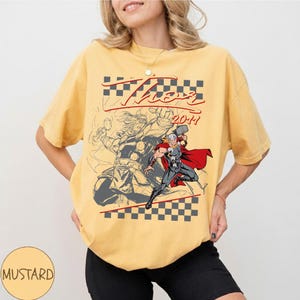 May include: Mustard-colored t-shirt featuring a graphic of Thor with the text "Thor 2011". The design includes a checkered pattern and a sketch-like illustration of Thor wielding his hammer. The shirt has short sleeves.