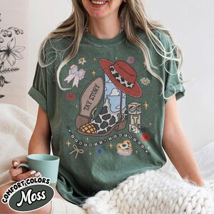 May include: A moss green short-sleeve t-shirt featuring a graphic print of a cow-print cowboy boot, a red hat, blocks with the letters T, S, and a star, and the text TAY STORY. The design includes small flowers, a bow, and beaded strings.