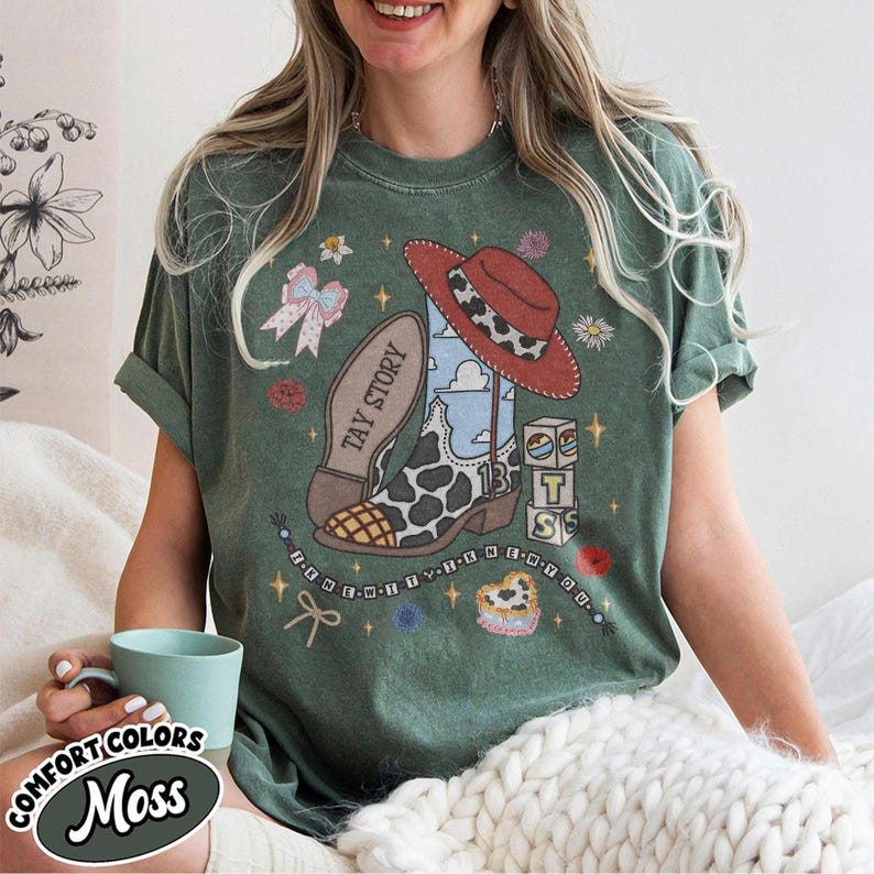 May include: A moss green short-sleeve t-shirt featuring a graphic print of a cow-print cowboy boot, a red hat, blocks with the letters T, S, and a star, and the text TAY STORY. The design includes small flowers, a bow, and beaded strings.