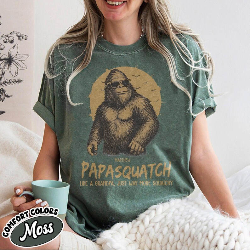 May include: Moss green t-shirt featuring a Bigfoot graphic wearing sunglasses in front of a yellow sun. The text "PAPASQUATCH" is below the graphic, with the phrase "LIKE A GRANDPA, JUST WAY MORE SQUATCHY". The shirt is part of the Comfort Colors collection.